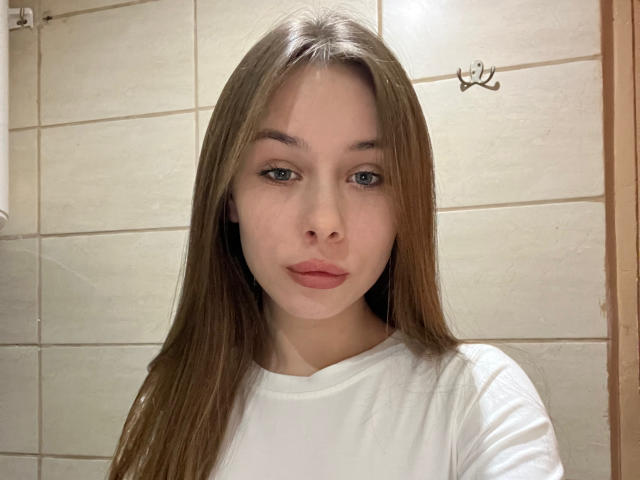 ElsaSexxy has long brown hair, blue eyes, and pink lipstick. She's wearing a white shirt, looking straight at the camera with a neutral expression.