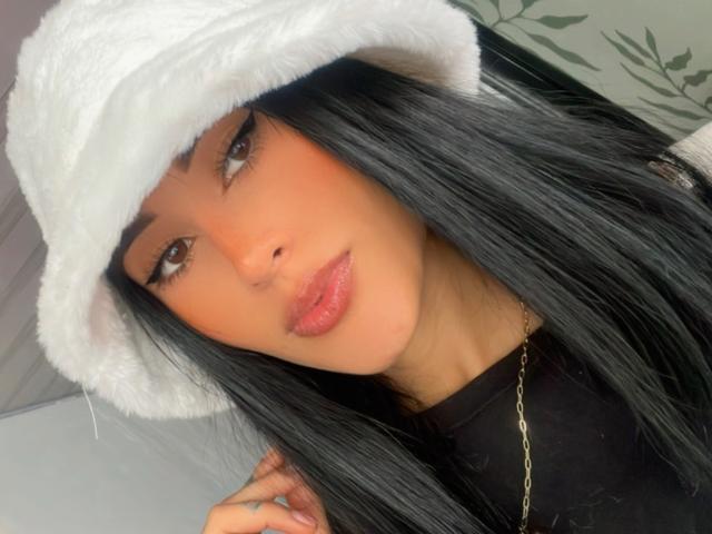 HarleyRoods has long black hair, brown eyes, and pink glossy lips. She's wearing a white fluffy hat and a black top with a gold chain.