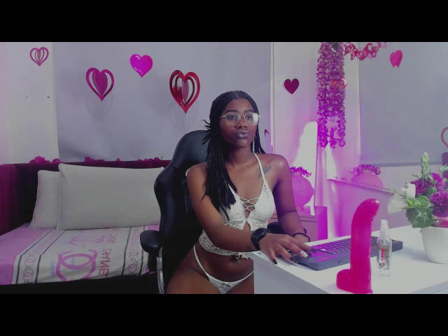 AriannaGreyy with dark skin, wearing a white lace bra and panties, sitting at a desk with a pink dildo, computer, and flowers. She has glasses and braided hair.
