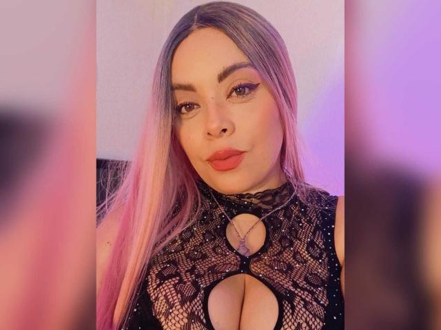 VictoriaRoyale with long pink-tinted hair, wearing a black lace top with a keyhole cutout showing her large, shiny tits. She has full lips, makeup, and a confident expression.