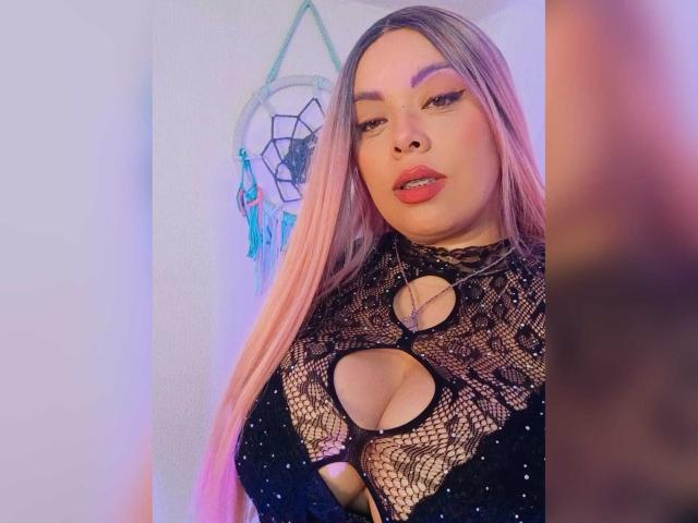 VictoriaRoyale with pink hair, full lips, and black lace top showing her big, cleavage-filled tits. She's looking at the camera with a slightly open mouth.