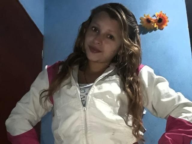 KeiliSexy has long brown wavy hair, wearing a white and pink jacket, standing with hands on hips, smiling slightly, with a flower decoration on the blue wall behind.
