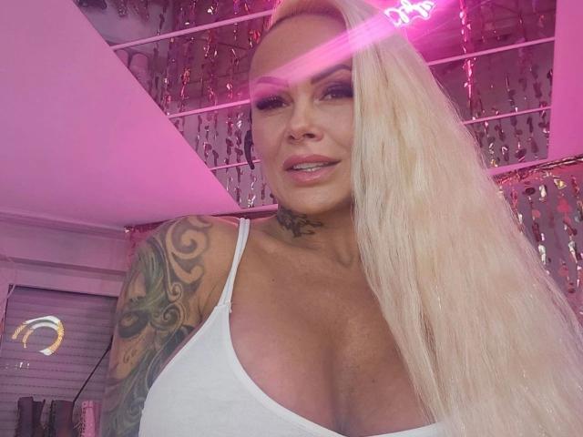 BonnievonClyde-hot with long blonde hair, white tank top, tattooed arm, and large tits, smiling under pink neon lights, showing a sexy, confident look.