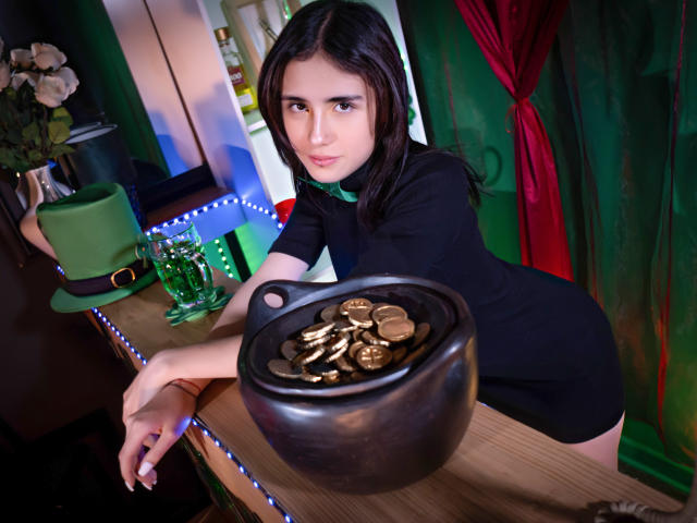 ValerieBanks with dark hair, black dress, leaning on a bar, holding a black pot filled with coins, green hat, and glasses in the background, red curtain to the right.