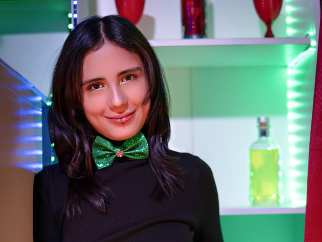 ValerieBanks with dark hair, green glittery bowtie, smiling, wearing black top, neon green and blue light background, red and green decor, yellow bottle in shelf.
