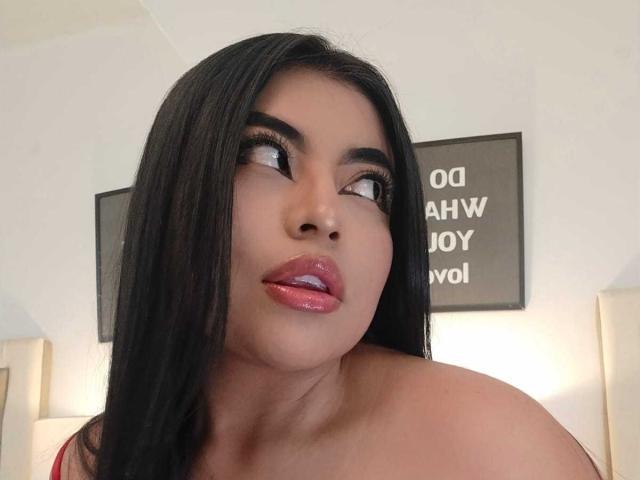 AlexaMadisson with shiny black hair, looking up with glossy pink lips, bare shoulders, and dark eyebrows, in a selfie. Her skin is tan, and she's wearing red lipstick.