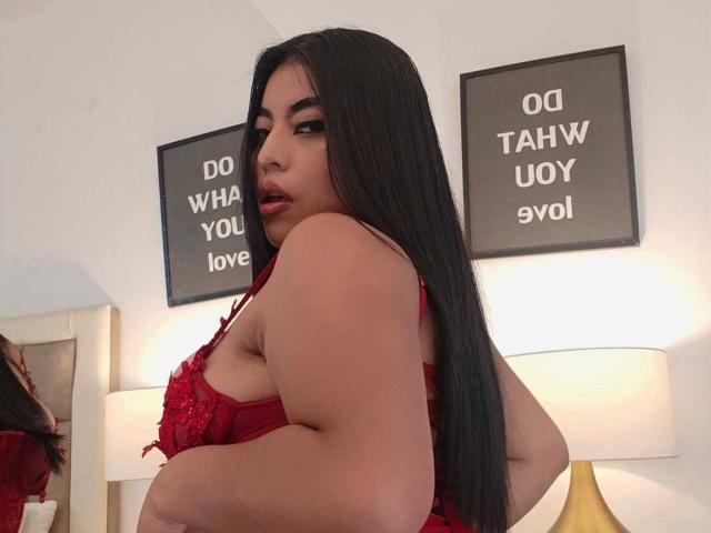 AlexaMadisson with long black hair, red lace lingerie, showing her tits, looking over her shoulder, red lipstick, white walls, and a gold lamp in the background.