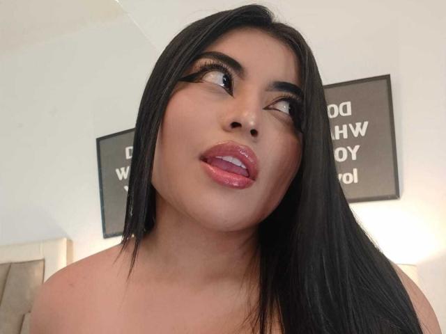 AlexaMadisson has long black hair, thick eyebrows, and shiny pink lips, looking up with her tongue slightly out. Her skin is tan, and she's wearing no top.