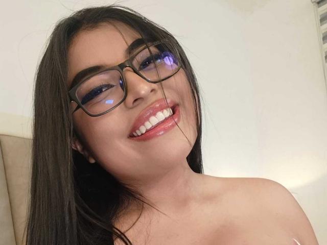 AlexaMadisson with glasses, smiling, showing shiny lips and white teeth, brown hair, and bare shoulders, looking cute and sexy.