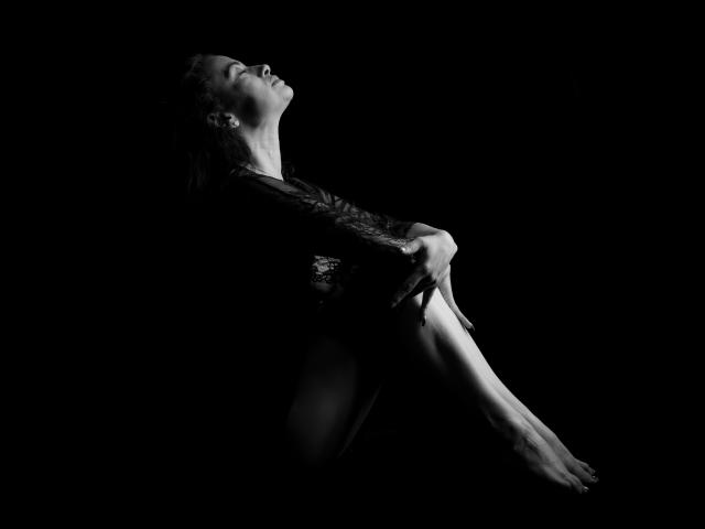AfroditaRgenis is curled up in a dark, shadowy space, her face tilted back, arms wrapped around her knees, bare feet visible, highlighting her tits and smooth skin.