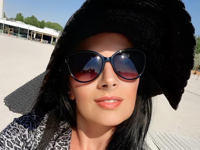 HannahFaith with black sunglasses, a large black sunhat, and a patterned shirt, showing her shiny pink lips and smooth skin. Her dark hair is slightly visible.