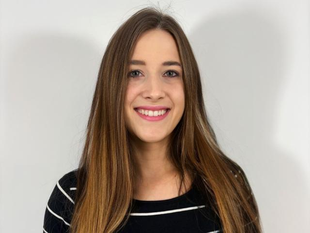 BellaSensitive69 with long brown hair, smiling, wearing a black striped shirt, has pink lipstick, and brown eyes, against a plain white background.