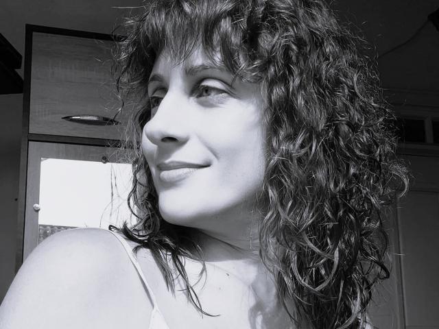 MarinaSexyAndHot with curly, wet-looking hair, smiling at the camera, showing her nose and lips, wearing a white, thin-strapped top, in a black-and-white photo.