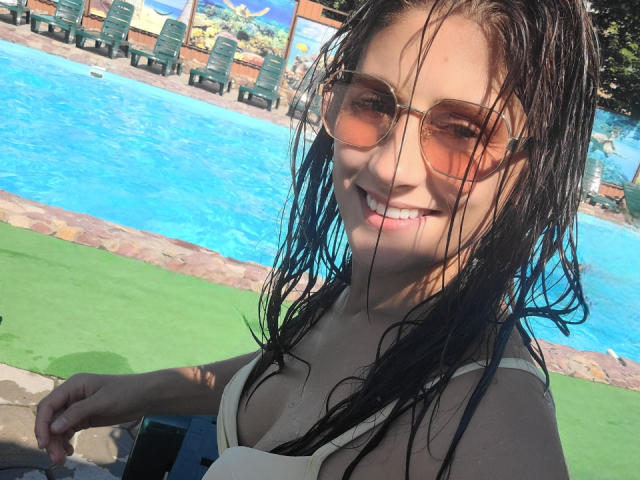MarinaSexyAndHot with wet black hair, smiling, wearing a white bikini top and aviator sunglasses, standing by a blue pool. Her tits are small, and she's holding a phone.