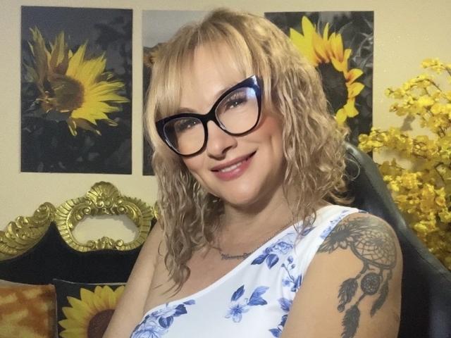 Cexy, con cabello rubio rizado, gafas negras, vestido blanco con estampado floral y un tatuaje en el brazo derecho, sonriendo. Pinturas de girasoles y flores amarillas en el fondo. Se ven los senos debajo del vestido..