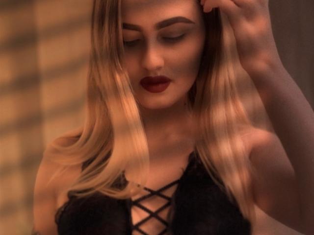 BabeCandy69 with blonde hair, wearing black lace-up lingerie, has red lipstick, and is looking down with her hand in her hair, showcasing her tits. Soft, warm lighting.