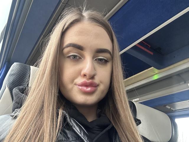 BabeCandy69 with long blonde hair, full pink lips, slight nose piercing, and blue eyes, wearing a black jacket, sitting in a bus, looking at the camera, smiling.