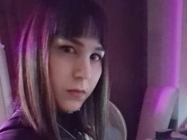 ShilaHot with straight black and purple-dyed hair, dark eyeliner, nose piercing, and lip piercing, looking slightly to the side with a serious expression. Purple and white lighting.