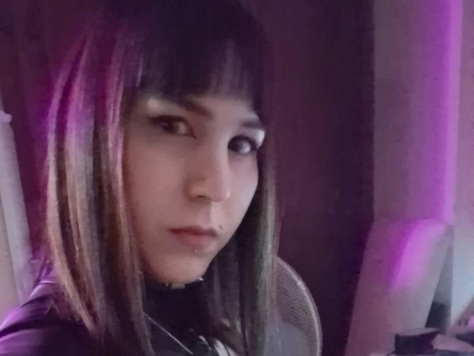 ShilaHot with straight black and purple-dyed hair, dark eyeliner, nose piercing, and lip piercing, looking slightly to the side with a serious expression. Purple and white lighting.