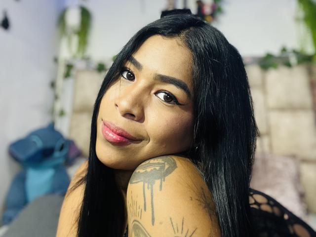MollyXDamiam with black hair, brown eyes, pink lipstick, and a tattooed shoulder, smiling at the camera, showing her tits slightly. She's on a bed with a blue bag.
