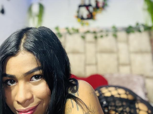 MollyXDamiam with black hair, brown eyes, and a tattoo on her shoulder, smiling at the camera, wearing a black, mesh, fishnet top, showing her tits.