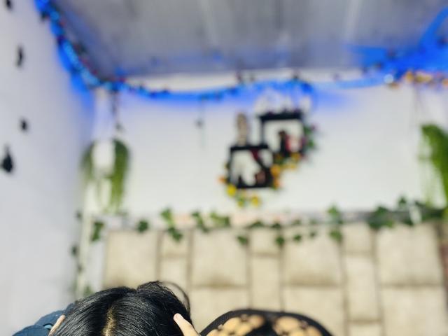 MollyXDamiam's black hair in foreground, back of head visible, looking at blurry wall with two framed photos and green leaf decorations, blue lights on ceiling.