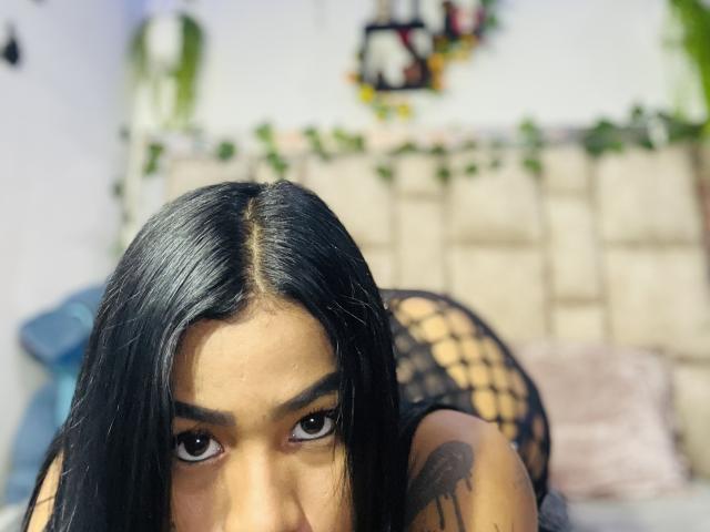 MollyXDamiam with black hair, looking up, in a fishnet dress, on a bed, showing a tattoo on her arm, and a focused expression. Her tits are slightly visible.