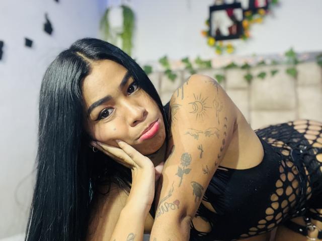 MollyXDamiam with long black hair, brown skin, and tattoos, wearing a black fishnet top, lying on a bed, head on hand, looking at camera with pink lips.