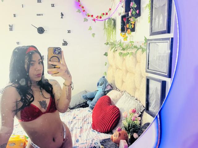 MollyXDamiam with long black hair, red bra, and thong, holding phone, showing tattoo on chest, bed with stuffed blue toy, heart pillow, and flowers in mirror selfie.