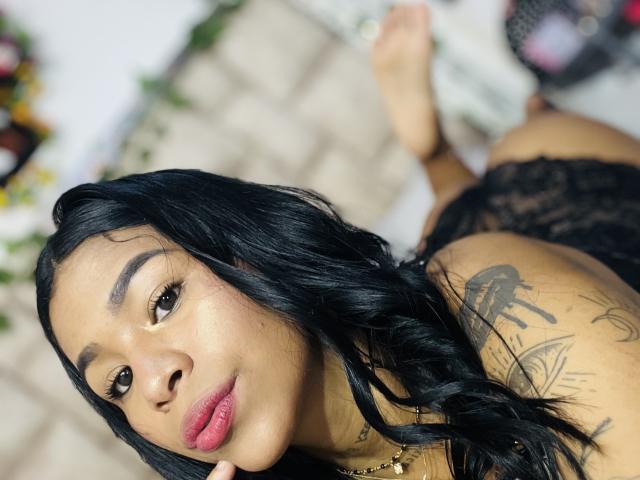 MollyXDamiam with black wavy hair, tan skin, tattoos, wearing lace lingerie, lying on her stomach, looking at the camera, showing her pouty pink lips and soft brown eyes.