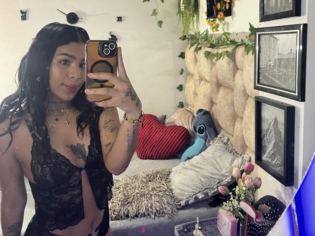 MollyXDamiam with black lace lingerie, showing her tits and tattoos, taking a mirror selfie with her phone. She's got long black hair and a slight smile.