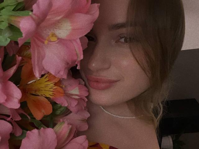 AlisaRiden's face with pink flowers, showing off her tits slightly, brown hair, pink lips, and a pearl necklace, in a dimly lit photo.