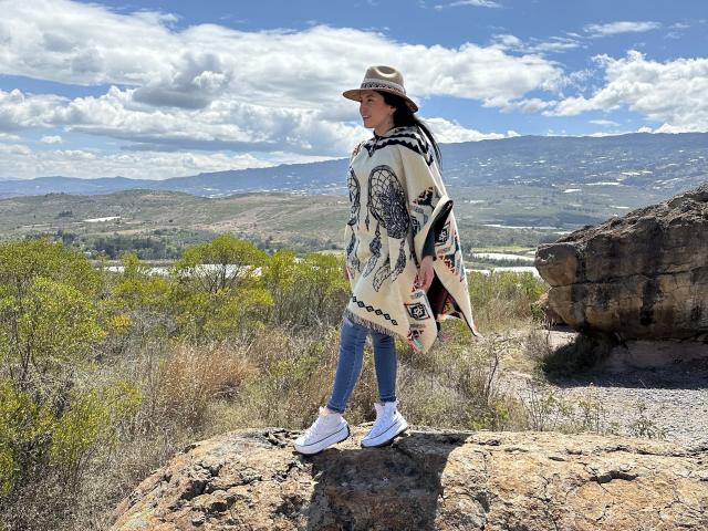 AmelieMuller stands on a rock, wearing a tan hat, black and white patterned shawl, skinny jeans, and white sneakers, with a mountain landscape and cloudy sky in the background.