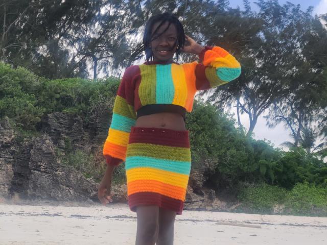 KrisJohanas with dark skin, smiling, wearing a colorful striped crop top and skirt, standing on a sandy beach, hand in hair, trees in background.