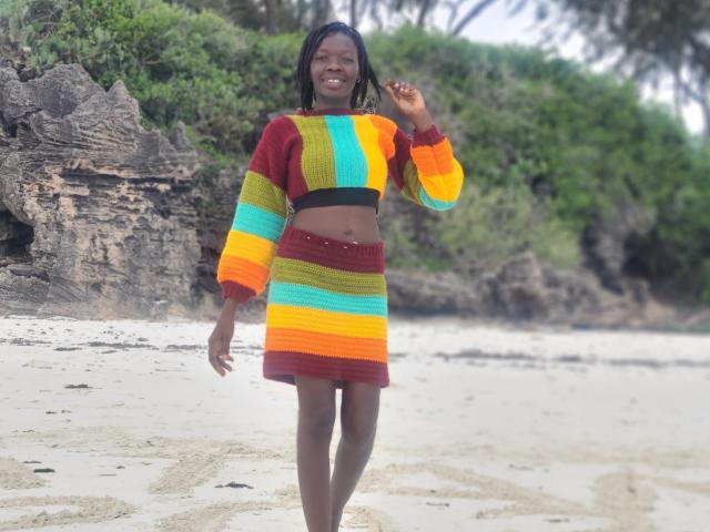 KrisJohanas with dark skin, black hair, wearing a colorful striped sweater and skirt, smiling, waving on a sandy beach with rocky cliffs and greenery in the background.