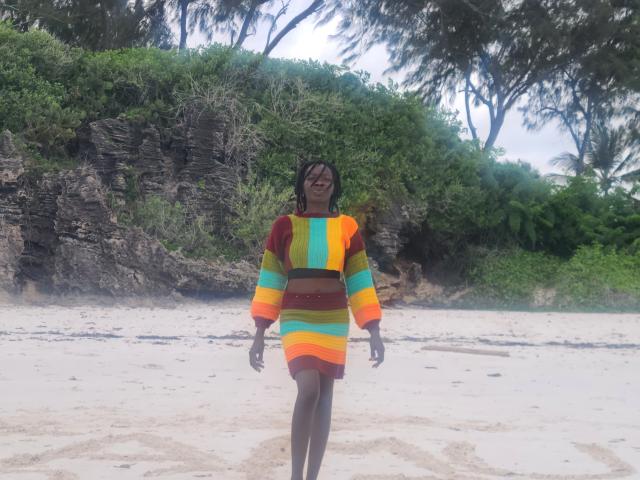 KrisJohanas with dark skin, standing on a sandy beach, wearing a colorful striped long-sleeve dress, smiling, and walking with arms slightly out. Her ass is small.