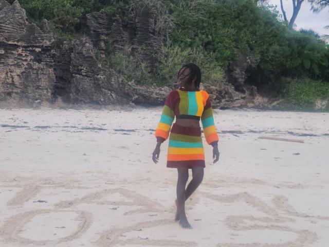 KrisJohanas wearing a colorful, striped dress with orange, green, yellow, and red patterns, standing on a sandy beach, facing away, brown skin, black hair, barefoot.