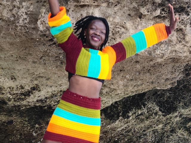 KrisJohanas with dark skin, smiling, arms raised, wearing a colorful striped knit crop top and skirt, exposing a black bra, standing against a rocky wall.