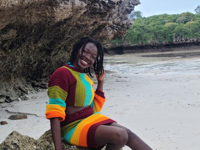 KrisJohanas, dark-skinned woman with dreadlocks, smiling, wearing a colorful striped knit crop top and skirt, sitting on a rock, beach background, colorful clothes, happy, relaxed.