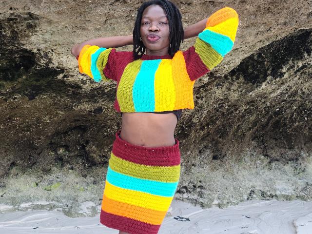 KrisJohanas with dark skin, medium build, and black dreadlocks, wearing a yellow, teal, and red striped knit crop top and skirt, hands behind head, standing on sandy beach.