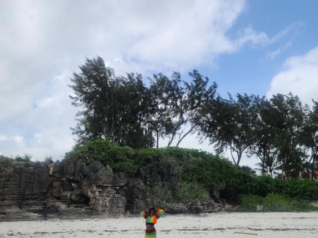 KrisJohanas stands on sandy beach, arms raised, wearing colorful clothes, facing rocky cliff with tall trees and green shrubbery under a partly cloudy blue sky.