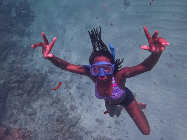 KrisJohanas with dark skin, wearing a purple bikini, blue snorkel mask, and blue headband, underwater with arms spread, black dreadlocks, and small orange fish near her.