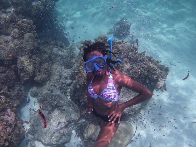 KrisJohanas with dark skin, wearing a blue-patterned bikini top, black bottoms, blue snorkel mask, and black snorkel, underwater with rocks and small fish around.
