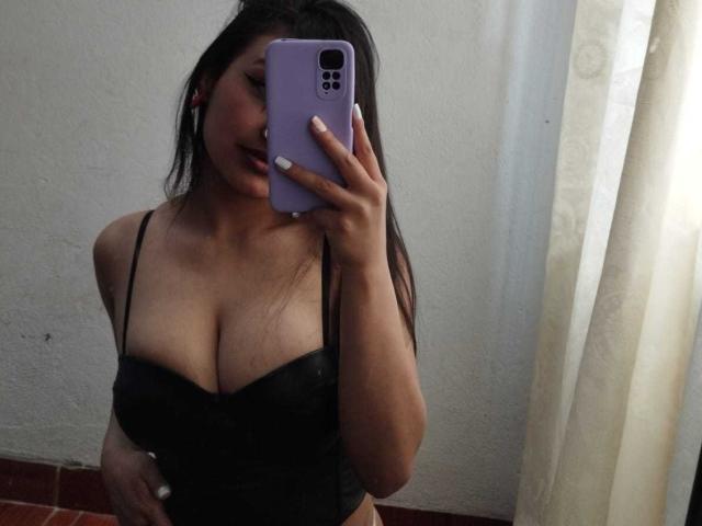 Elisa Turner, con un seno di media grandezza, indossa un top nero senza spalline e sta scattando un selfie allo specchio con un telefono viola, ha i capelli lunghi e scuri, le unghie bianche e un leggero sorriso..