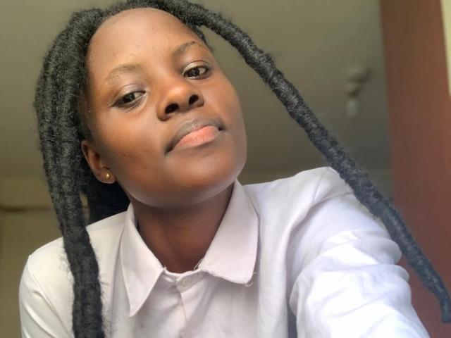 SexyBadriya with dark skin and black dreadlocks, wearing a white shirt, has a subtle smile and small gold earring, looking slightly up at the camera.