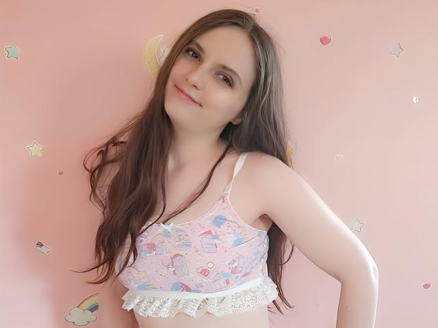 SweetJulhia with long brown hair, wearing a pink and blue patterned bra with lace trim, smiling softly at the camera, showing her small tits and cute face.