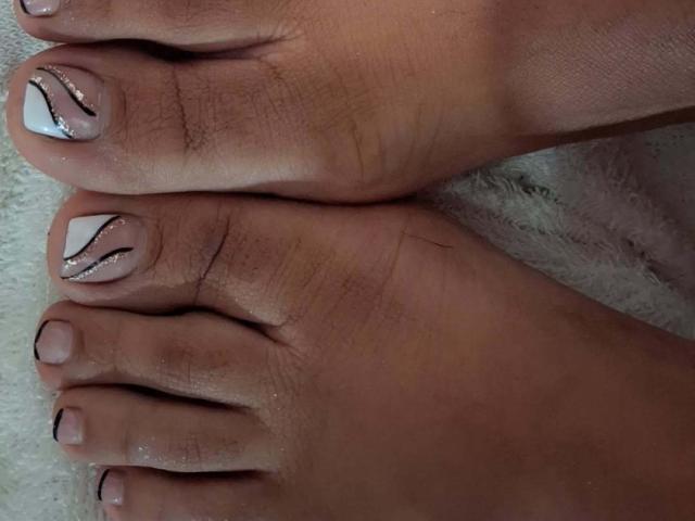 JackelineRoset's feet, showing her toenails with white nail polish and black, wavy, silver-trimmed designs. Her toes are slightly dirty with visible skin texture.
