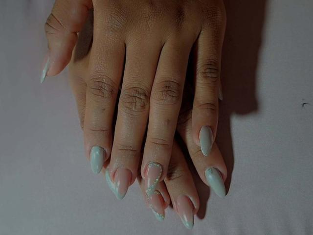La main de Jackeline Roset, avec de longs ongles pointus de couleur rose pâle, chacun orné de petites paillettes vertes, repose sur une surface blanche, montrant des articulations détaillées et des doigts propres et soignés..