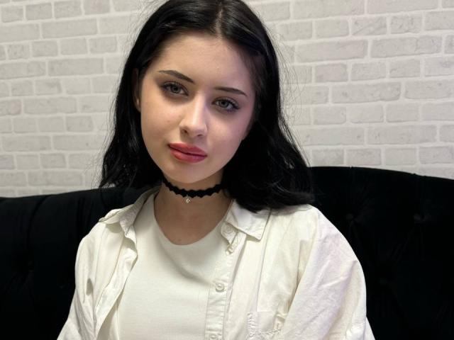 LovelDiva with black hair, wearing a white shirt, black choker, and pink lipstick, has a cute face and small tits, sitting on a black couch.