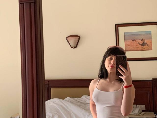 LovelDiva with black hair, wearing a white tank top, showing her small tits, holding a phone, standing in a bedroom, looking at the mirror, red wristband.
